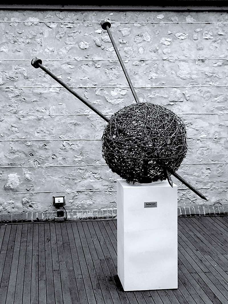 Black and white image showing a round sculpture with knitting needles in it.