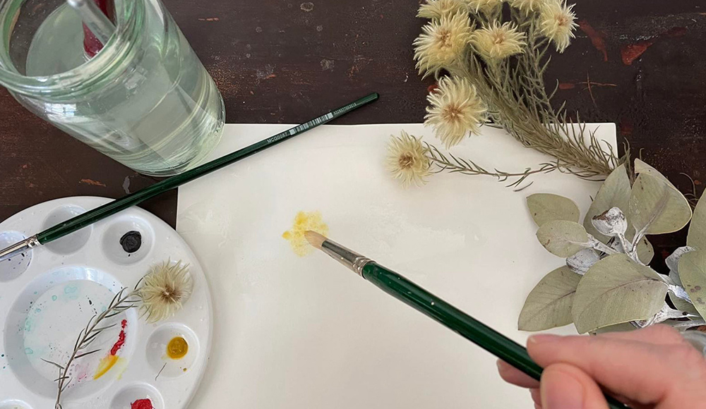 Image shows a hand holding a paintbrush painting a yellow flower on a white page. To the right are some dried flowers and gum leaves. To the left is a paint pallet and a jar of water.
